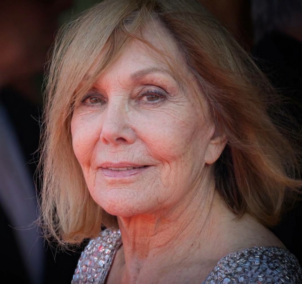 En un primer plano, Kim Novak lleva una chaqueta con detalles plateados y esboza una sutil sonrisa, mientras que una cálida iluminación resalta su rostro y su melena rubia. | Fuente: Gemini