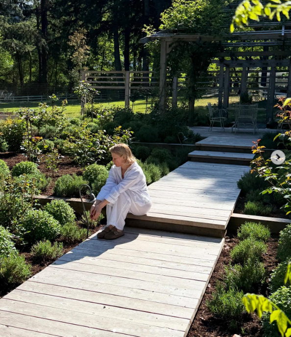 Pamela Anderson descansa en los escalones de un paseo de madera blanqueada en Arcady, sentada descalza con una holgada ropa de lino blanca entre arbustos de boj bien cuidados y parterres en flor. Con la mirada puesta en la vegetación frente a una pérgola de madera que da sombra, transmite la tranquila satisfacción de la autonomía, y acompaña la publicación con el siguiente texto: "Pienso en la vida como un jardín… Puedes volver a plantar, empezar de nuevo y hacerla tuya. 🤍 Con cariño, P". | Fuente: Instagram/pamelaanderson