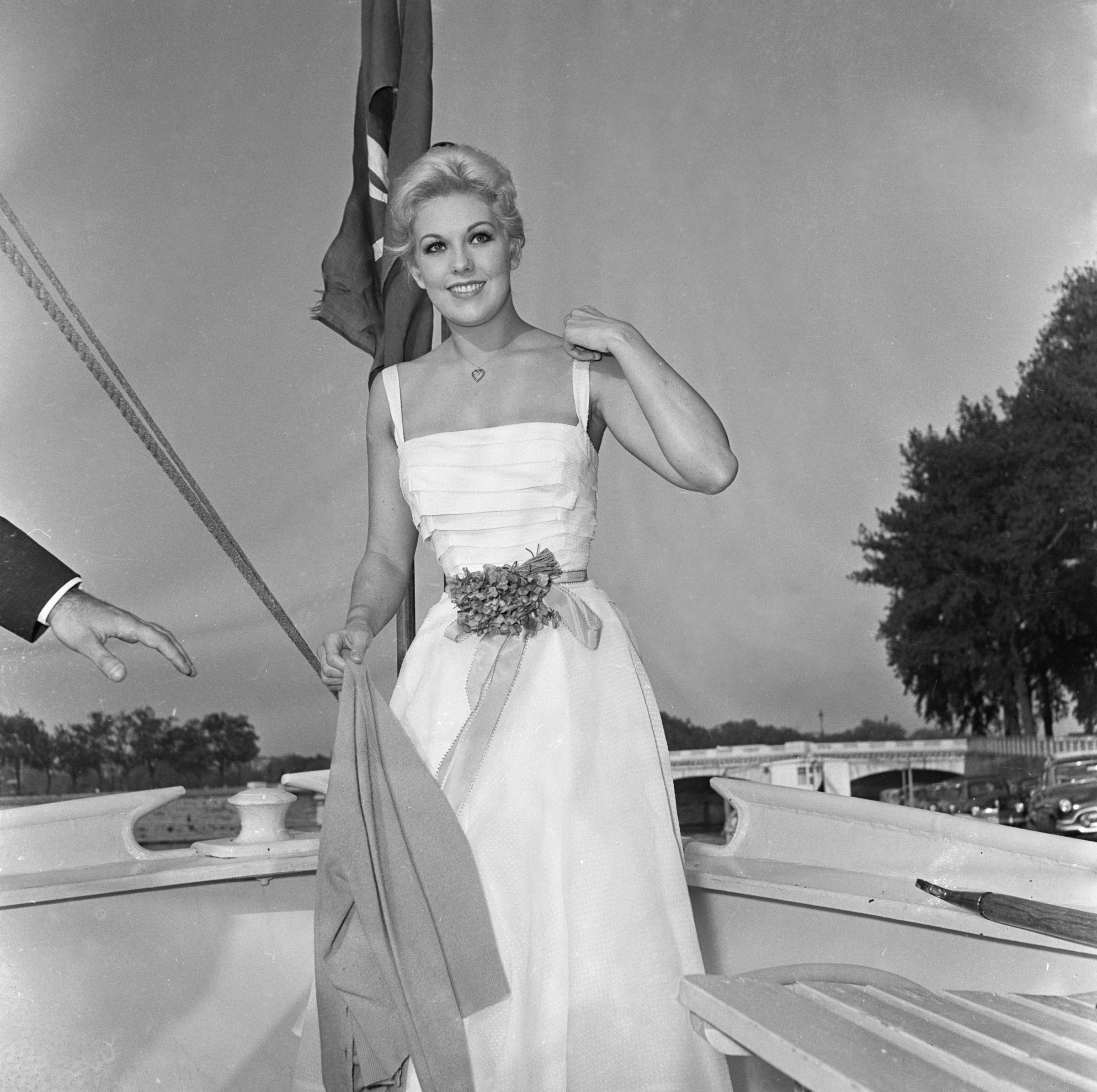 Kim Novak posa con un vestido blanco sin mangas a bordo del Borde-Brétigny, en el Sena, en París, sonriendo junto a una bandera. | Fuente: Getty Images