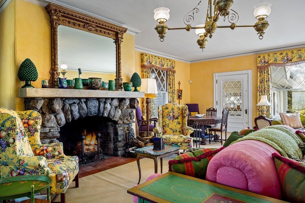 Una sala de estar muy colorida combina una gran chimenea de piedra con sillones con motivos florales, muebles de colores vivos y un espejo ornamentado sobre la repisa de la chimenea. | Fuente: Facebook/LegacySIR