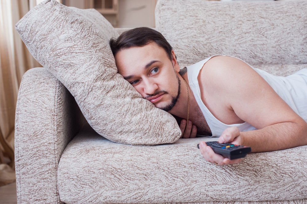 Hombre boca abajo en un sofá cambiando los canales de la televisión. | Fuente: Shutterstock