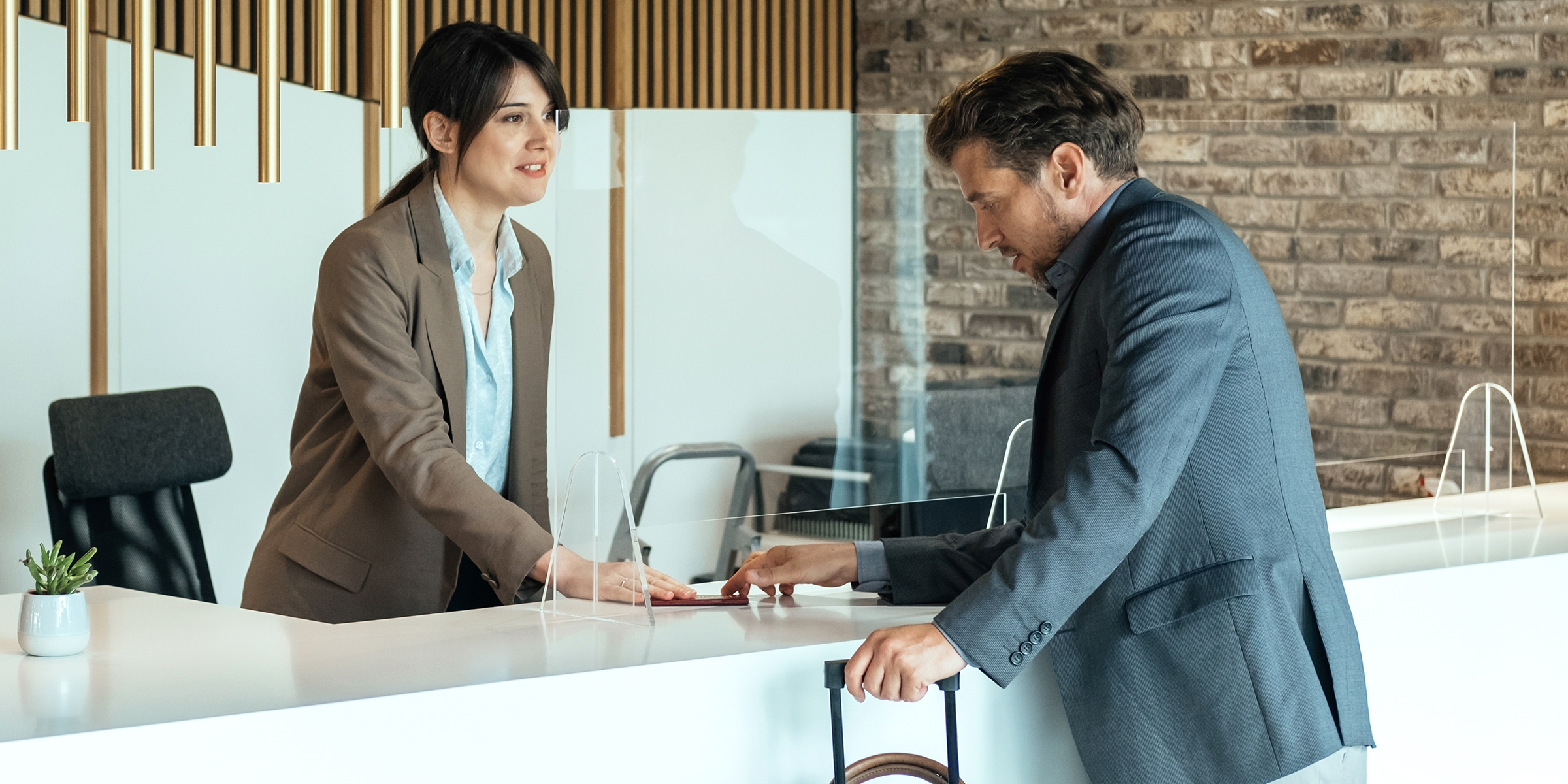 Un hombre se registra en la recepción de un hotel y habla con un recepcionista a través de un cristal protector | Fuente: Shutterstock
