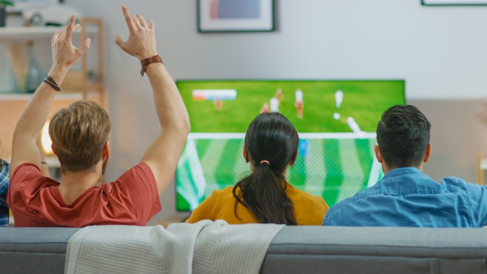 En casa, varios hinchas deportivos sentados en un sofá viendo un importante partido de fútbol en la televisión. I Foto: Shutterstock
