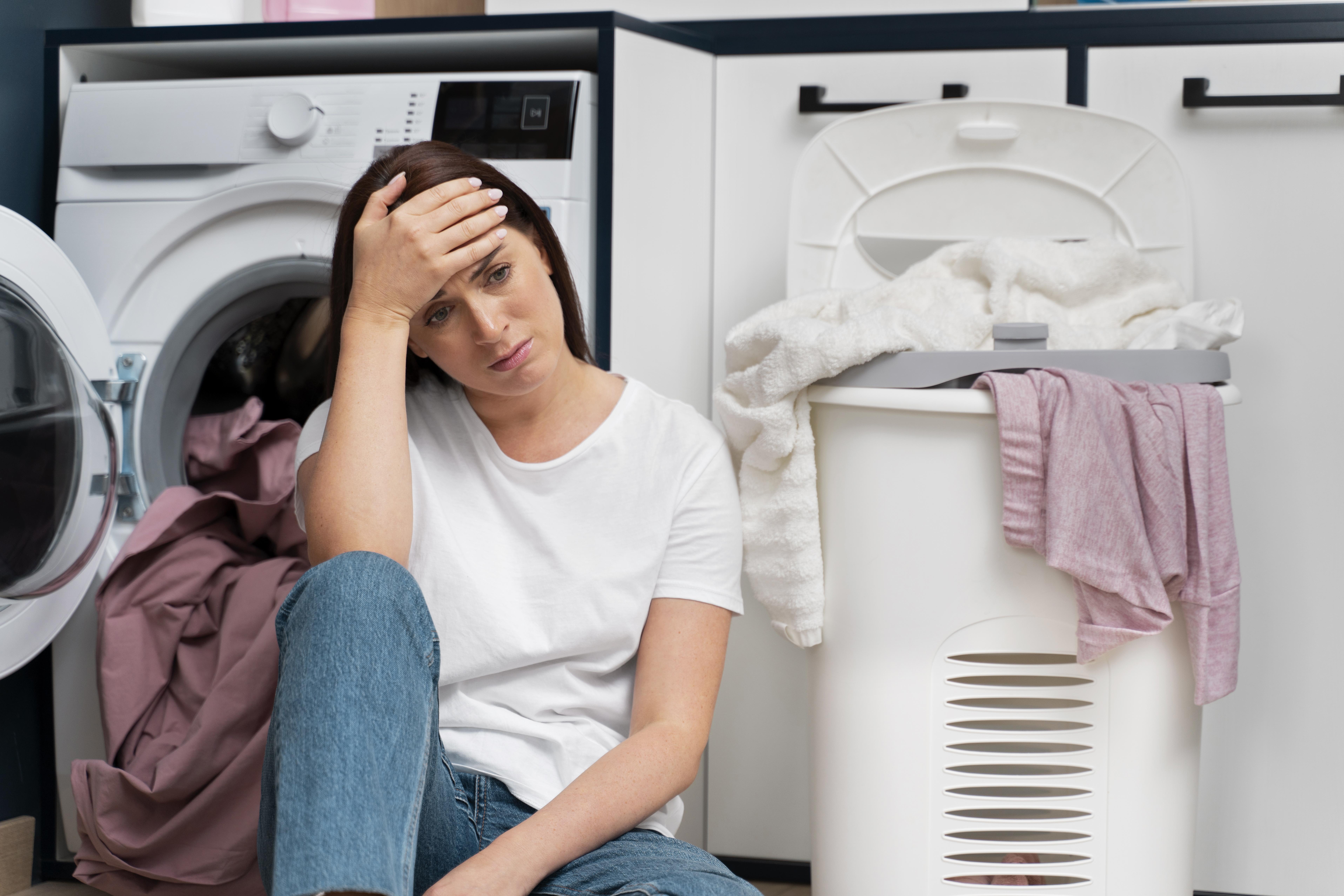 Mujer con cara de disgusto mientras se apoya en una lavadora, rodeada de ropa sucia | Fuente: Freepik
