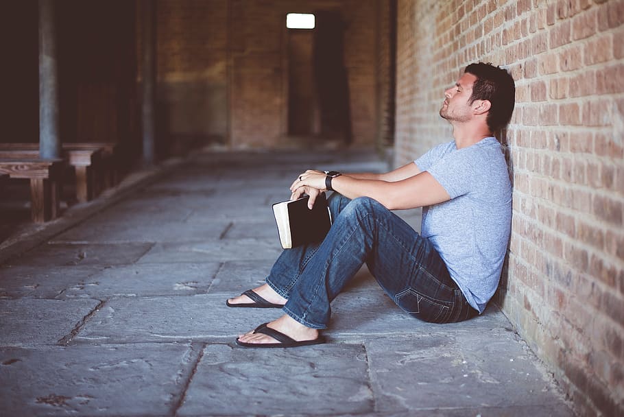Hombre pensativo sentado en el suelo recostado de una pared con un libro en sus manos. | Foto: PxFuel