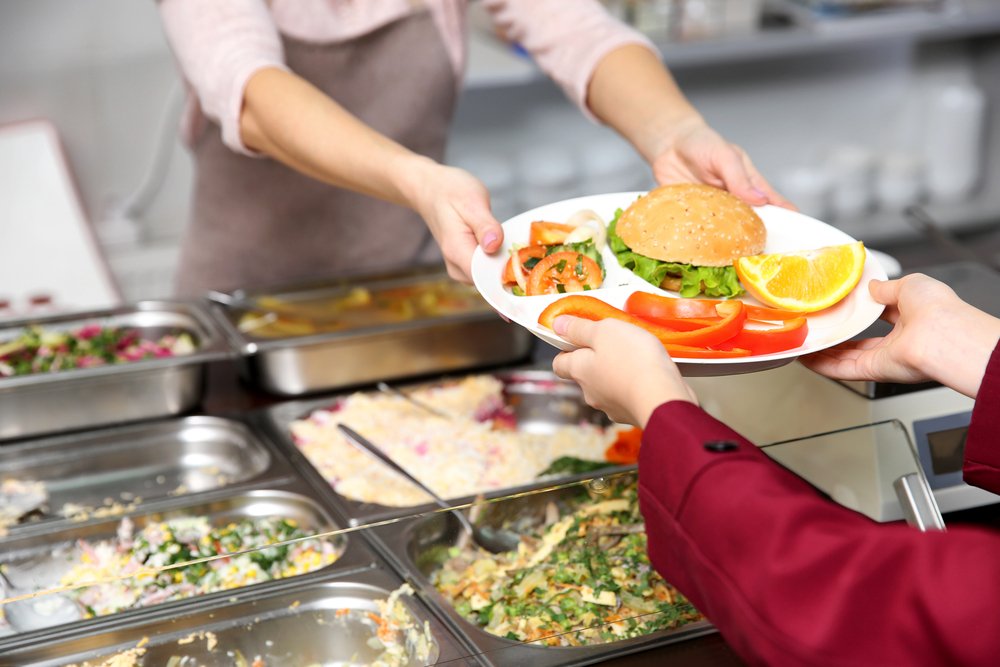 Mujer agradable dando almuerzo a una colegiala en la cafetería escolar. | Fuente: Shutterstock.
