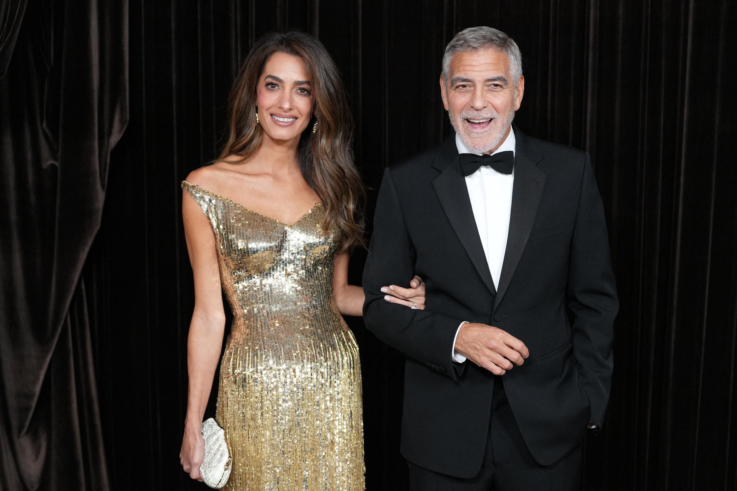 George y Amal Clooney posan cogidos del brazo en los premios Albies 2026, celebrados en Londres el 9 de octubre. Amal está espectacular con un vestido dorado brillante con hombros al aire y un bolso de mano a juego, mientras que George lleva un esmoquin negro clásico con pajarita. | Fuente: Getty Images