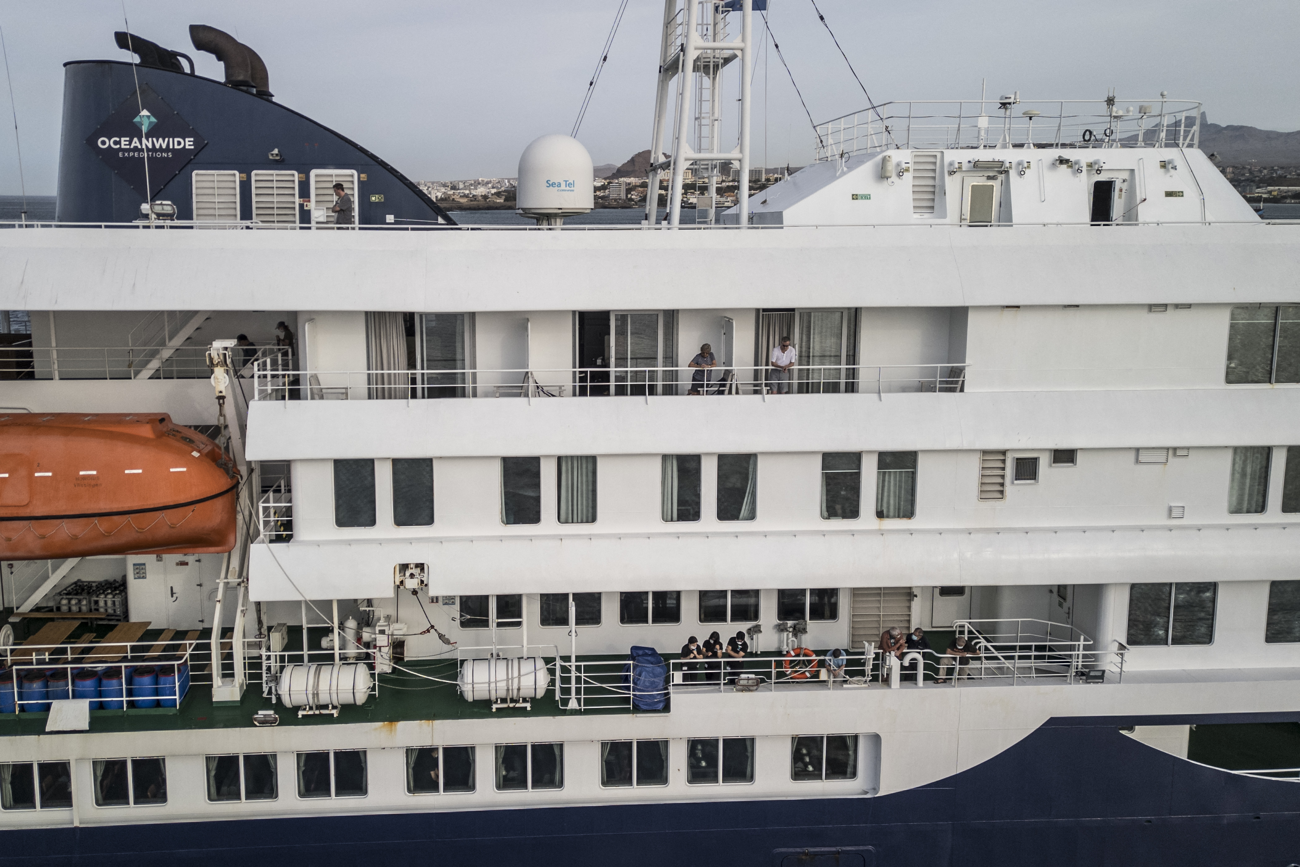 Esta vista aérea muestra a los pasajeros observando cómo personal sanitario, invisible para el espectador, ayuda a los pacientes a subir a un barco desde el crucero MV Hondius, mientras este se encuentra atracado frente al puerto de Praia, la capital de Cabo Verde, el 6 de mayo de 2026. | Fuente: Getty Images