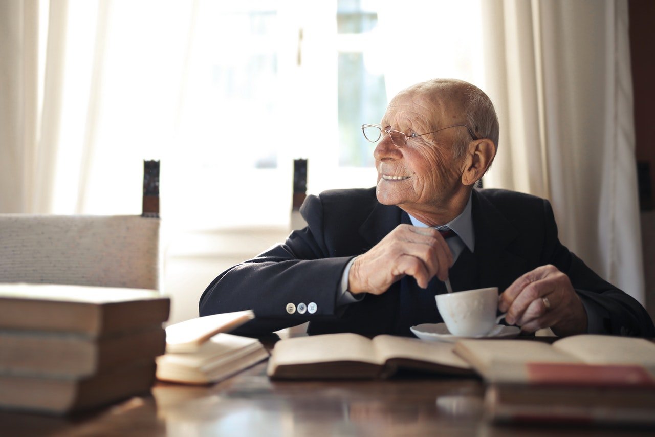 Anciano sonriente tomando una taza de té frente a un escritorio. | Foto: Pexels