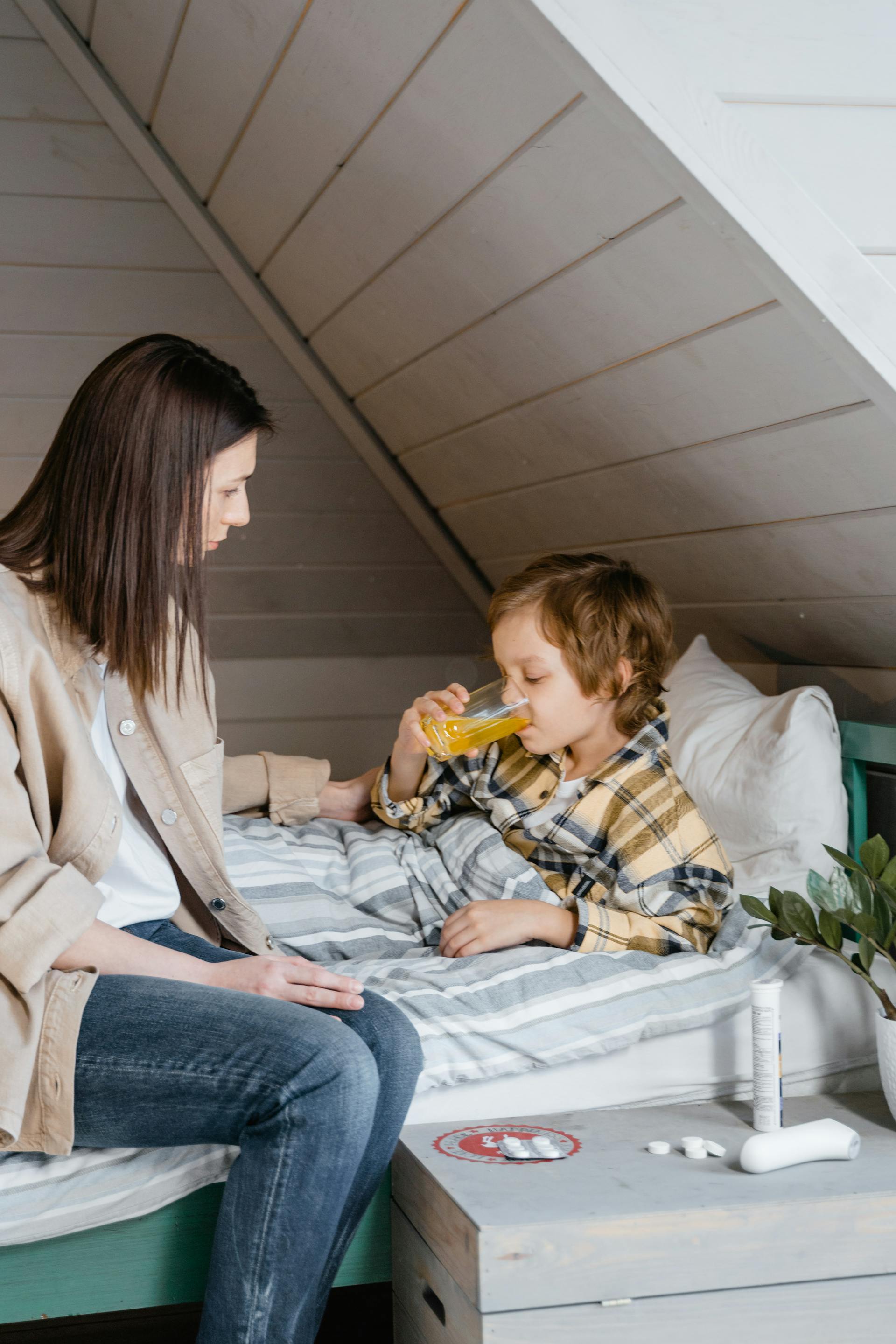 Una mujer mirando a su hijo pequeño bebiendo zumo en la cama | Fuente: Pexels