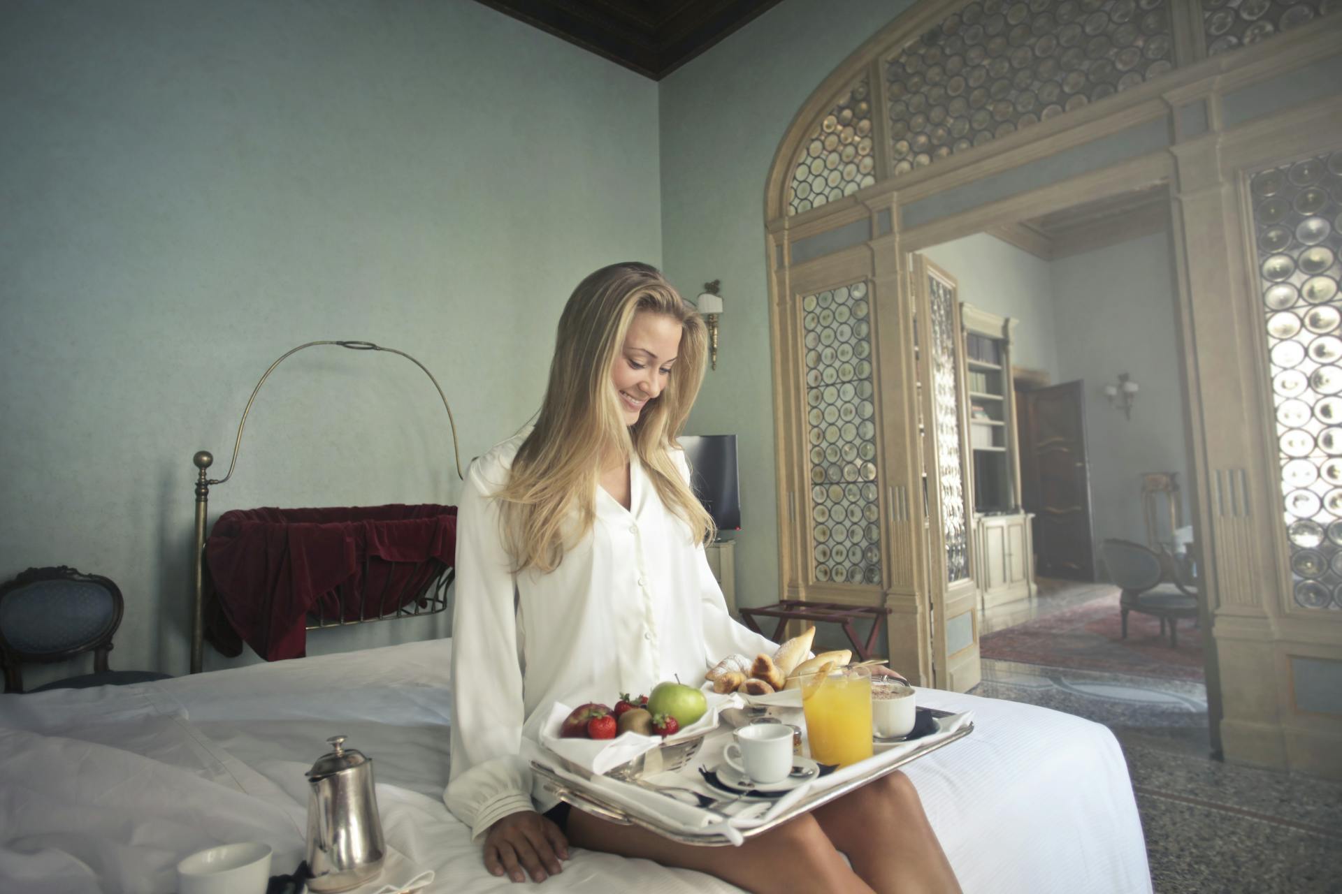 Una mujer alegre con el desayuno en una bandeja en la habitación de un hotel | Foto: Pexels