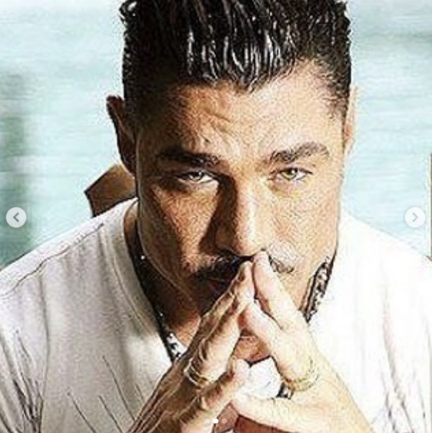 Ricardo Fort fue retratado en un plano cerrado con las manos juntas frente a su rostro, luciendo anillos y una cadena al cuello. La imagen captura su intensa mirada hacia la cámara, con el cabello peinado hacia atrás y una remera blanca clásica. | Fuente: Instagram/felipe_fort_