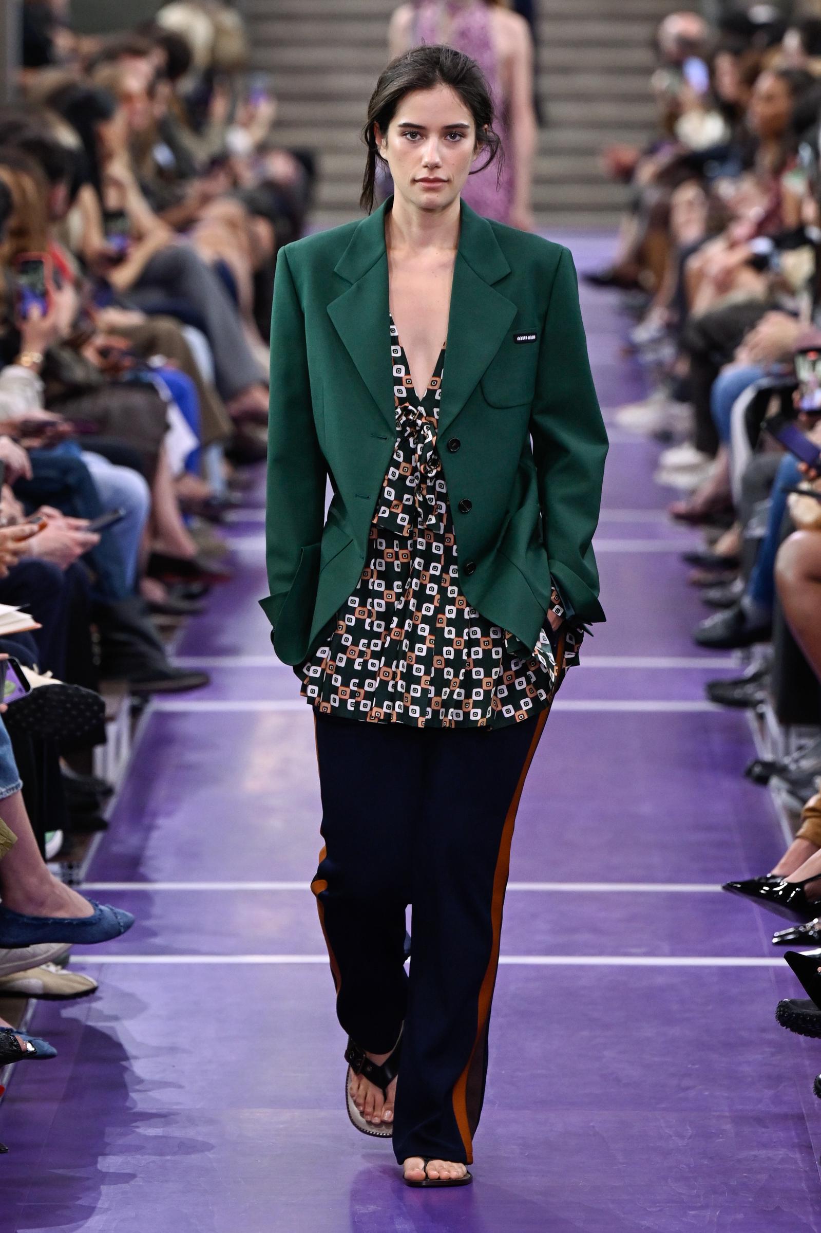 Ella Bleu Travolta hace su aparición en la pasarela de Miu Miu con una llamativa chaqueta verde sobre un top estampado, combinada con unos pantalones de chándal azul marino de corte holgado y unas sandalias planas negras. | Fuente: Getty Images