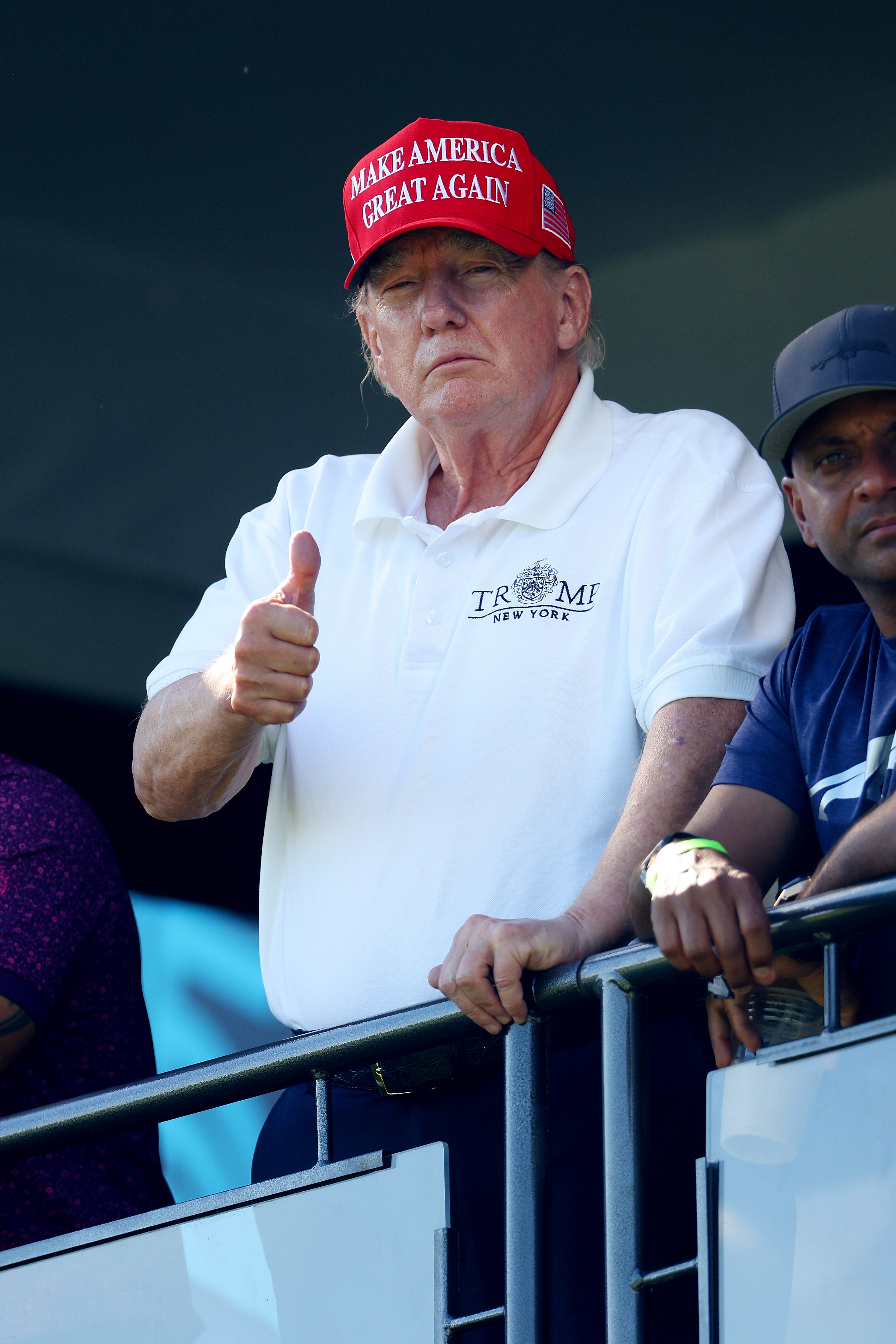 El 13 de agosto, Donald Trump está de pie cerca de una barandilla en el green del hoyo 18, dando un pulgar hacia arriba. El enrojecimiento de su rostro sigue siendo visible, con tonos más claros en los bordes de la mandíbula y el cuello.