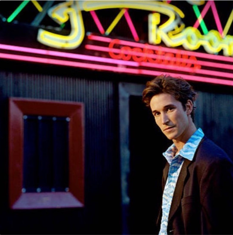 El actor, cuando era un joven adulto, posa frente a un local iluminado con luces de neón en una foto antigua de sus primeros años en Hollywood, con una camisa estampada y una chaqueta oscura. | Fuente: Instagram/therealnoahwyle