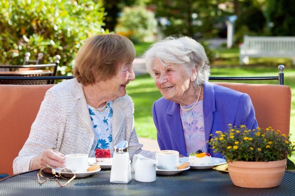 Dos mujeres mayores felices sentadas, charlando en la mesa al aire libre con café y bocadillos. Fuente: Shutterstock