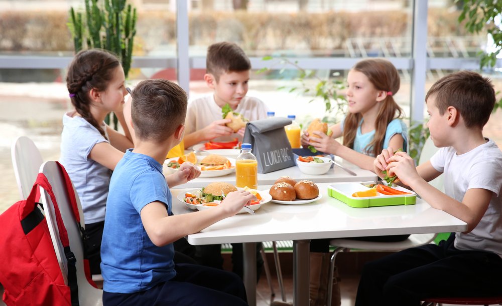 Niños sentados en la mesa de la cafetería mientras almuerzan. | Fuente: Shutterstock.