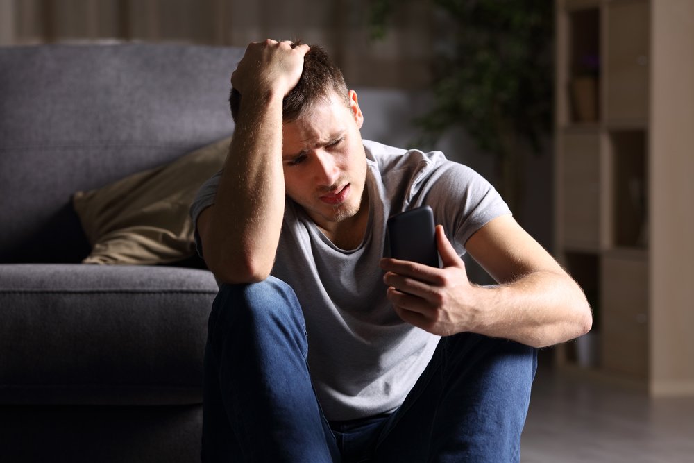 Hombre con rostro angustiado sentado en el suelo mientras sostiene un teléfono. | Foto: Shutterstock