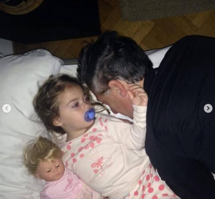 Antonia descansa acostada en la cama con su chupete y una muñeca al lado, vistiendo un pijama blanco con estampado rosa. A su lado, Mauricio Macri se recuesta sobre la almohada y le habla de cerca en un momento íntimo antes de dormir. | Fuente: Instagram/mauriciomacri