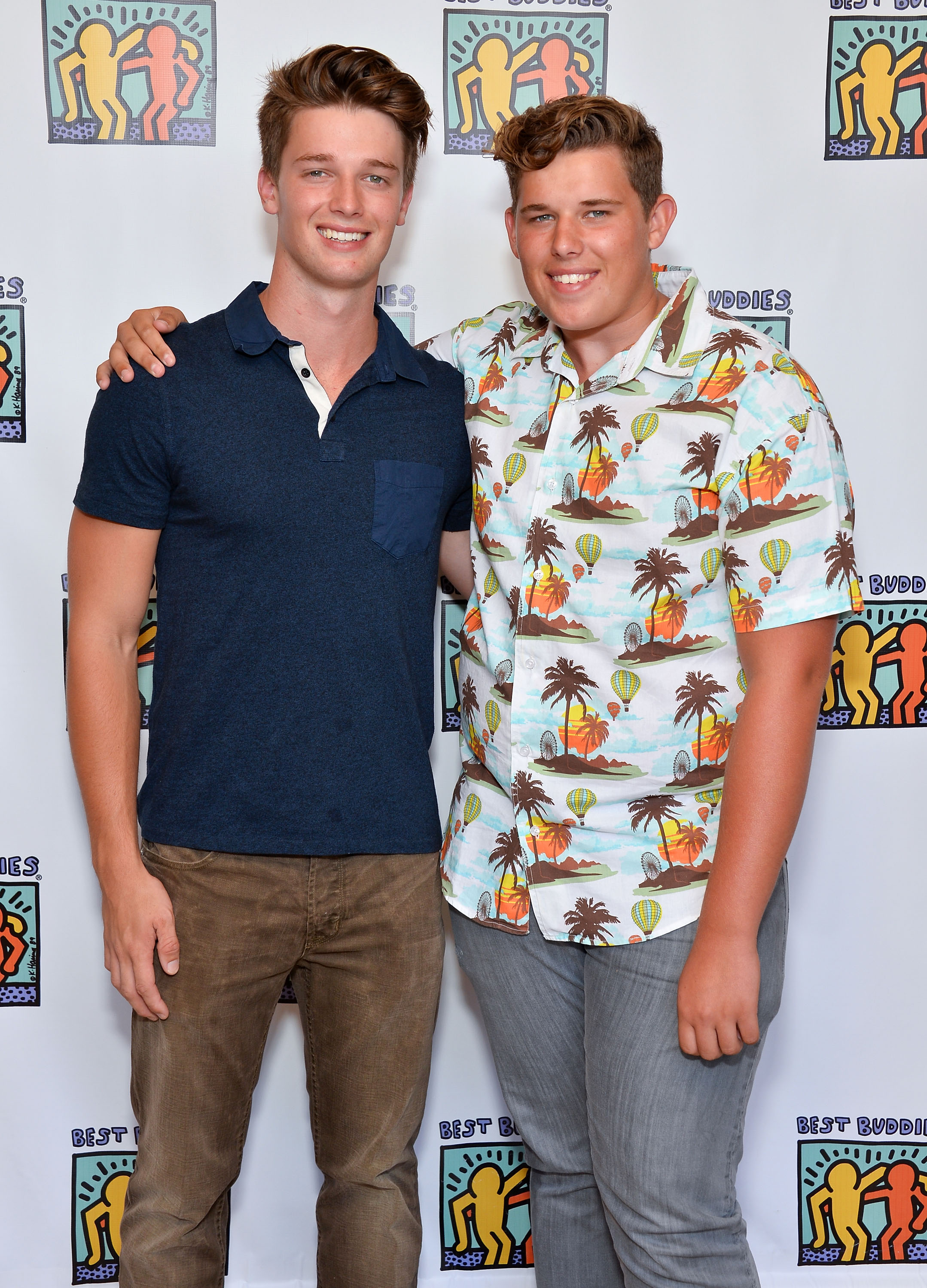 Patrick posa con su hermano pequeño Christopher en la gala benéfica del Equipo María para Best Buddies en Beverly Hills. Patrick va sencillo con una camisa de cuello azul marino y vaqueros marrones, mientras que Christopher destaca con una colorida camisa de botones de estampado tropical y pantalones grises, sonriendo con confianza a la cámara.