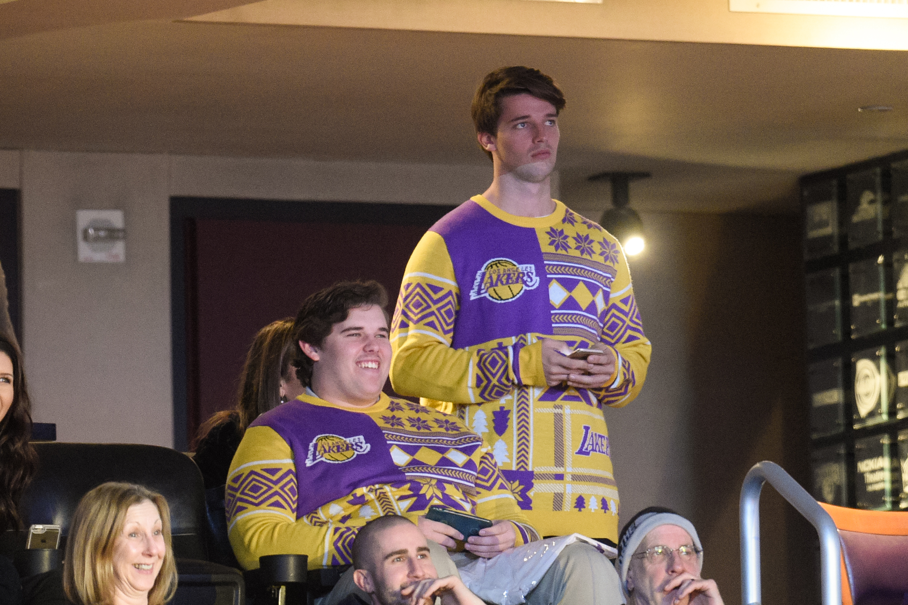 Christopher ve el partido de los Lakers con su hermano Patrick en el Staples Center de Los Ángeles en diciembre de 2015. Vestido con un atrevido jersey navideño de color morado y amarillo con temática de los Lakers, se ve a Christopher sentado y sonriente, con el pelo pulcramente peinado y la complexión notablemente más rellena en ese momento.