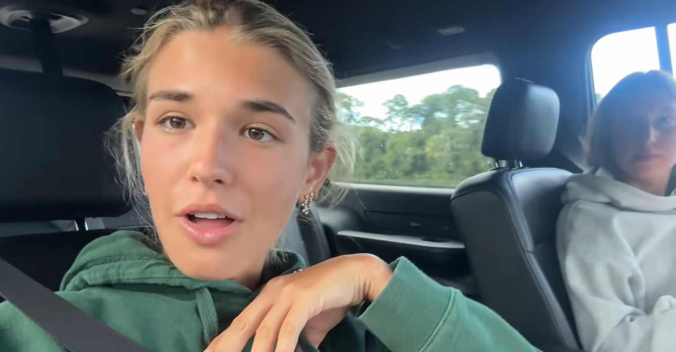 Kai Trump habla a la cámara desde un automóvil, vestida con una sudadera verde con capucha. Contó que había cogido faringitis estreptocócica durante su primera semana de clase y que había vuelto a Jupiter, Florida, para recuperarse con la ayuda de su madre. | Fuente: YouTube/kaitrump