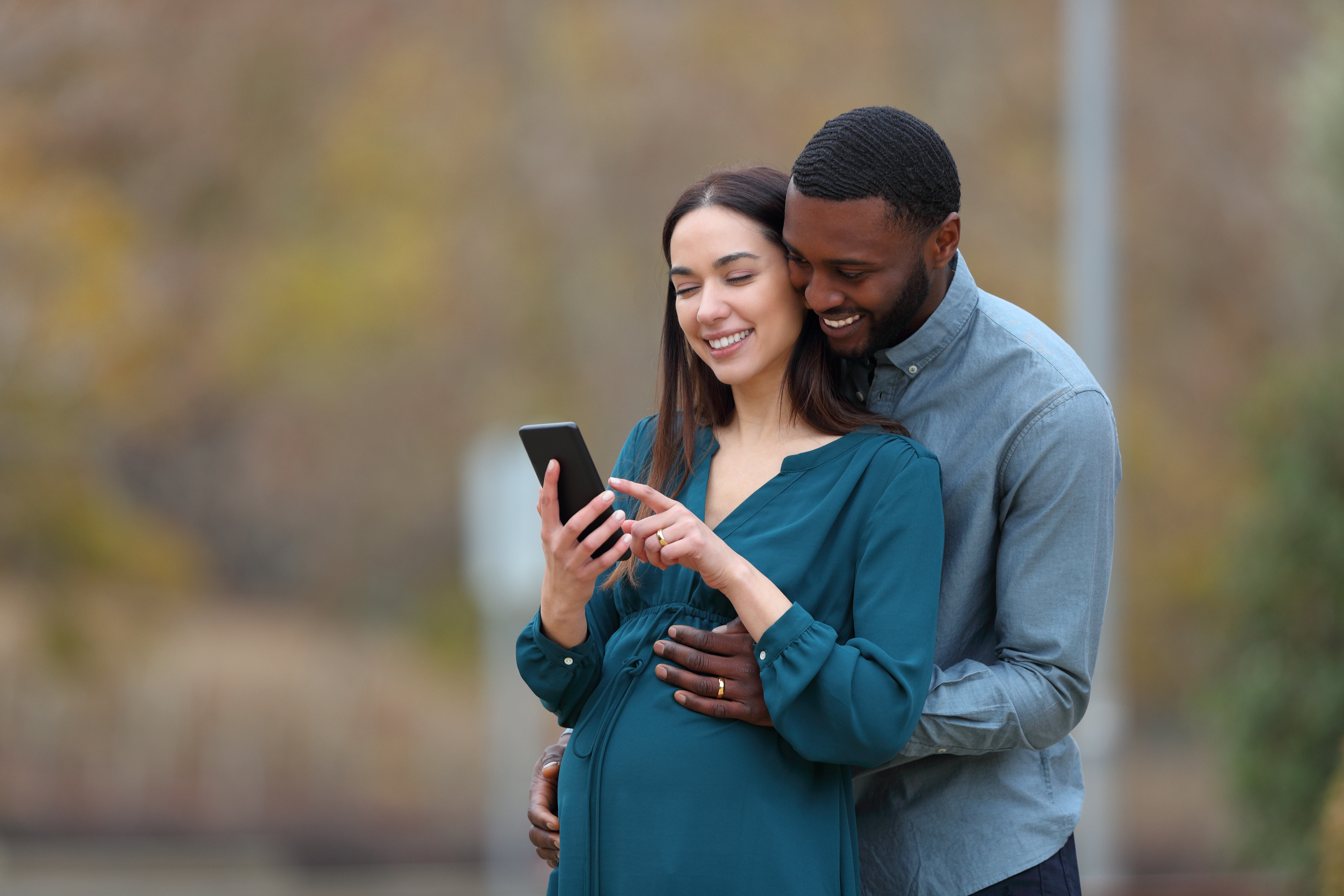 Una pareja interracial con una mujer embarazada y su marido consultando el smartphone en un parque | Foto: Shutterstock