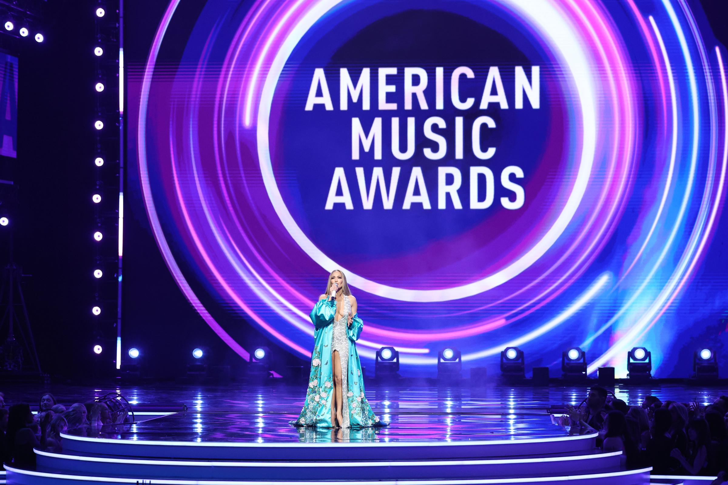 Sobre un vibrante remolino de rosas y azules, Jennifer Lopez cierra su momento en el escenario de los American Music Awards, erguida bajo el imponente logotipo del evento.