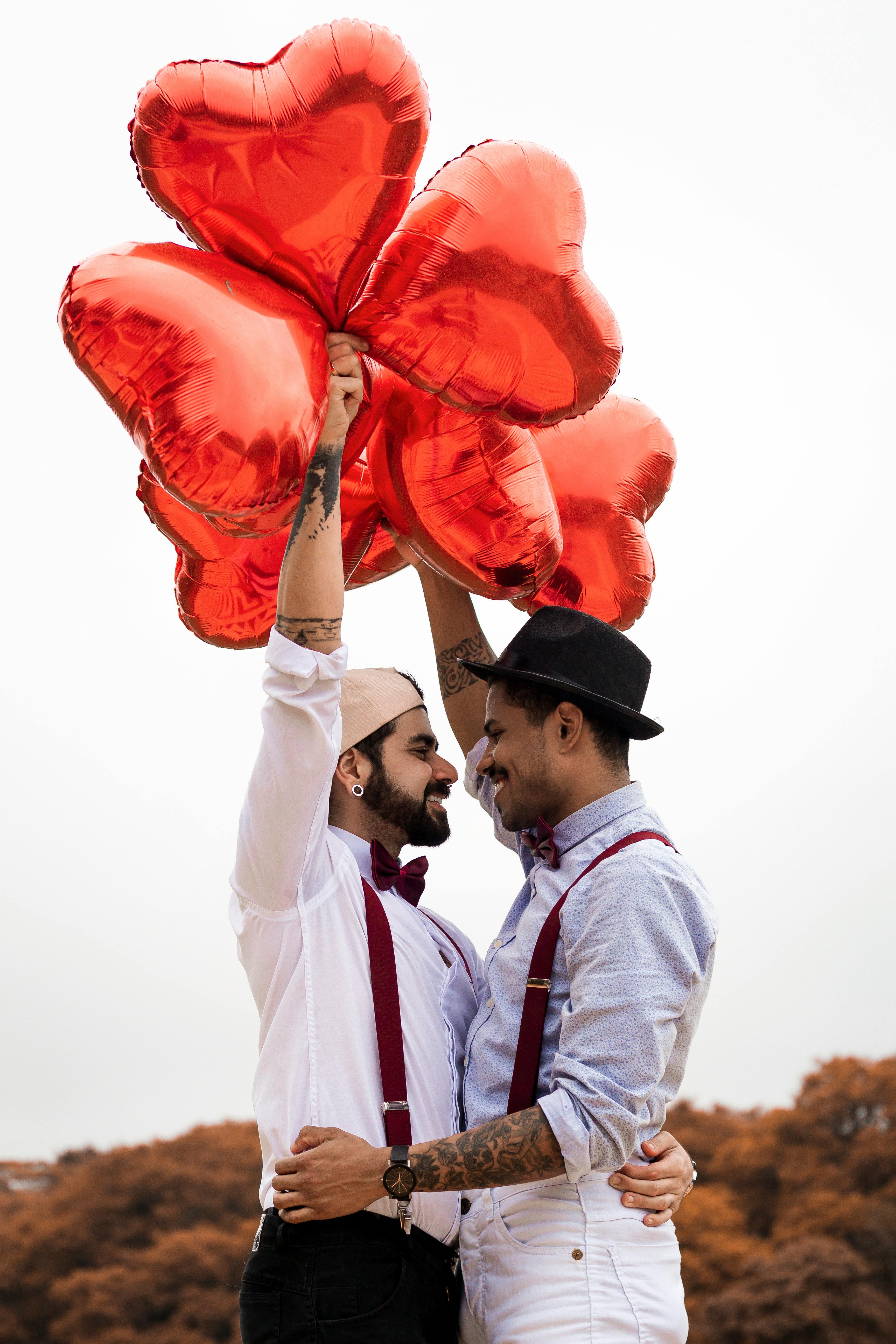 Dos hombres sonriendo y abrazándose mientras sostienen globos en forma de corazón | Foto: Pexels