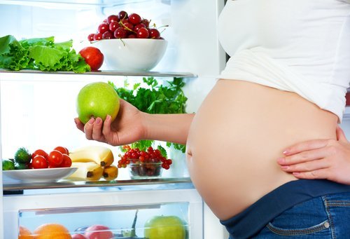 Mujer embarazada que se coloca cerca del refrigerador con las frutas y verduras. | Foto: Shutterstock