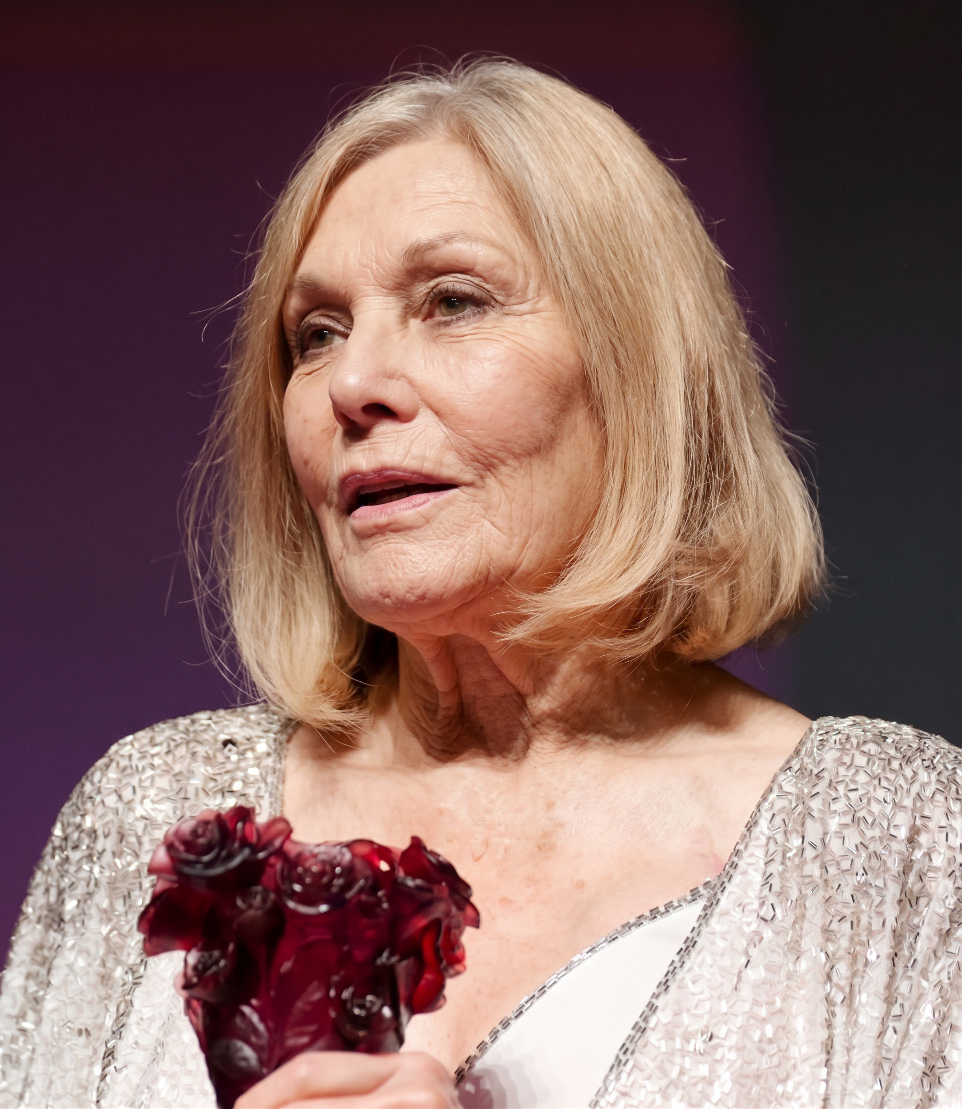 Kim Novak se gira ligeramente hacia un lado con una chaqueta plateada brillante y un vestido blanco, sosteniendo un premio con motivos florales de color rojo oscuro sobre un fondo morado apagado. | Fuente: Gemini