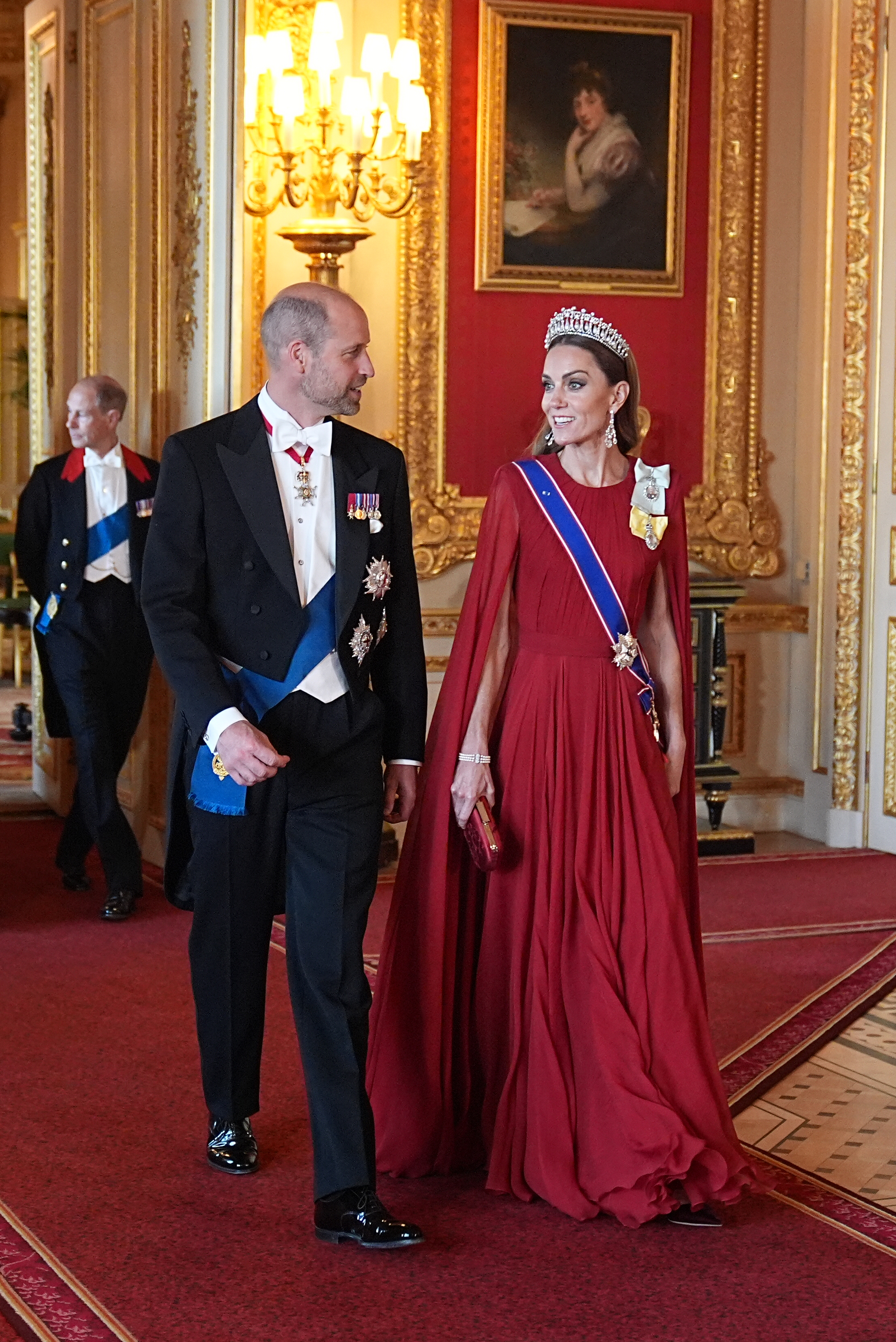 El príncipe William y Catherine, princesa de Gales, hacen una entrada triunfal al banquete de Estado en honor al presidente francés Emmanuel Macron y la primera dama Brigitte Macron, en el castillo de Windsor el 8 de julio de 2025. Catherine destaca con un vestido rojo carmesí plisado con capa, la tiara Lover's Knot de la reina Mary, pendientes colgantes de diamantes, la banda de la Real Orden Victoriana y la Orden de la Familia Real del rey Charles III. Sonríe junto a William, quien viste frac formal de Windsor, corbata blanca y condecoraciones ceremoniales. | Fuente: Getty Images