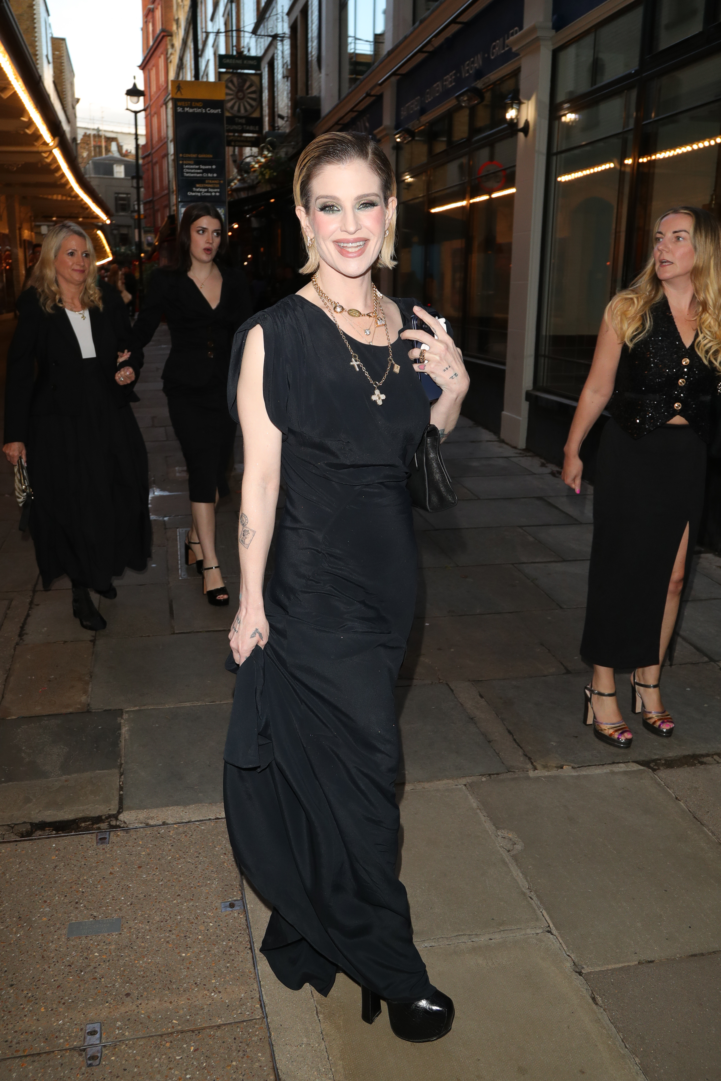 Kelly Osbourne sonrió a los fotógrafos durante una salida en Londres, con un vestido largo negro, varios collares cruzados y un peinado liso y peinado hacia atrás. | Fuente: Getty Images.