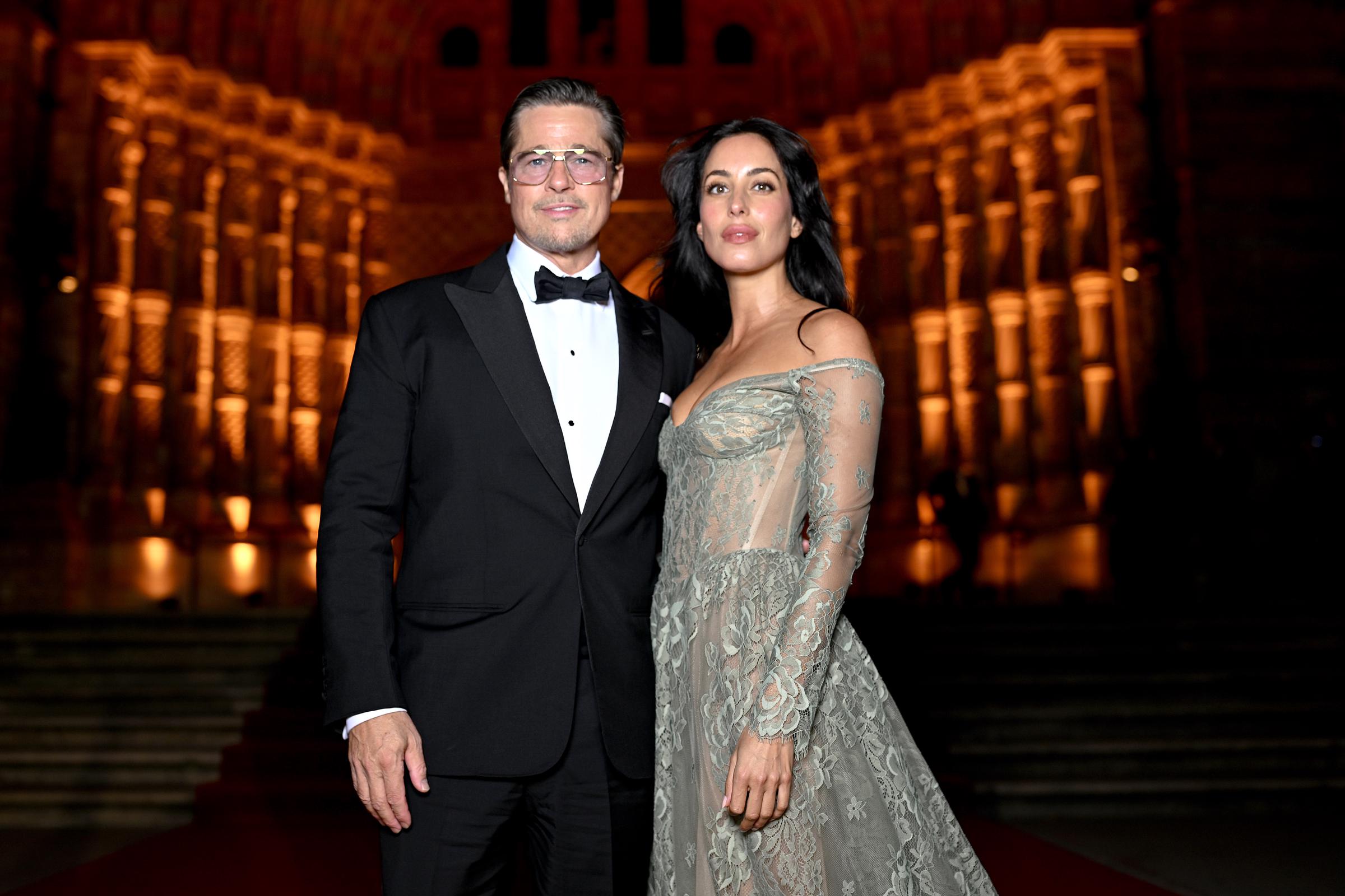 Brad Pitt e Inés de Ramón posan juntos en la escalera iluminada del Albies 2026 de Londres: Brad lleva un esmoquin negro y gafas de sol, e Inés luce un vestido de encaje transparente con hombros al aire, con intrincados bordados florales, un corsé ceñido y una cola fluida. Los delicados detalles del vestido y las mangas translúcidas destacan sobre la cálida iluminación dorada del lugar. | Fuente: Getty Images