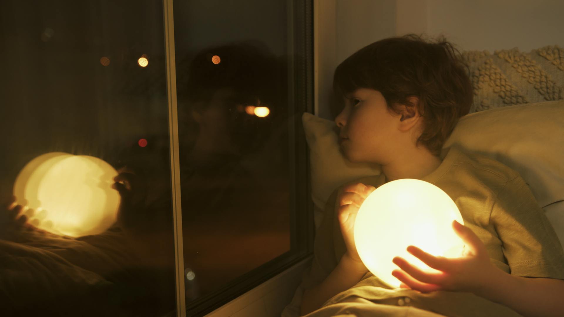 Un niño con una bola de luz asomándose por su ventana de noche | Fuente: Pexels