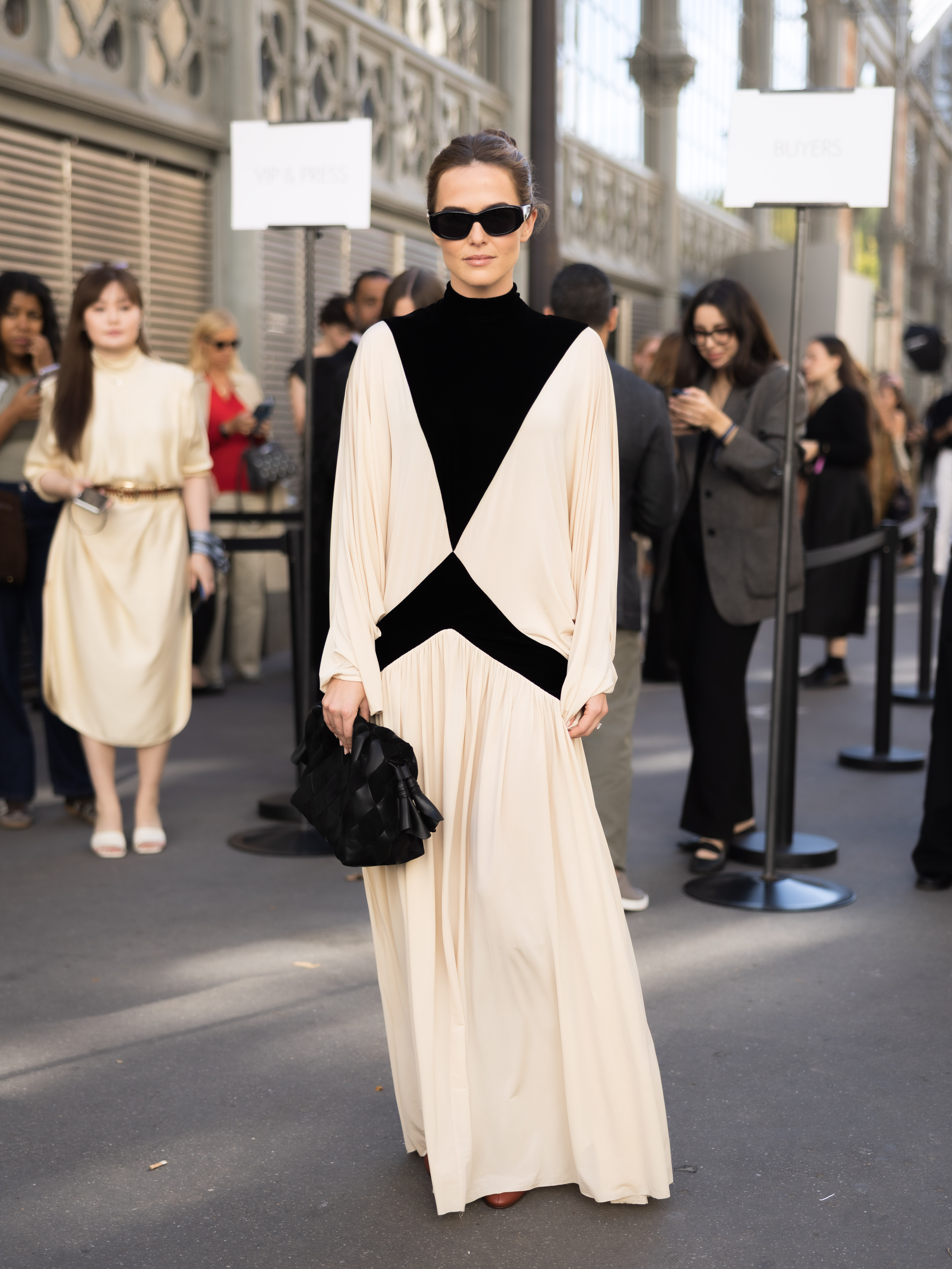 Unos llamativos paneles negros atraviesan el vestido color crema de Zoey Deutch, creando un marcado contraste geométrico desde el escote hasta la cintura. El resto del look lo mantiene sencillo, con unas gafas de sol oscuras y un bolso negro. | Fuente: Getty Images