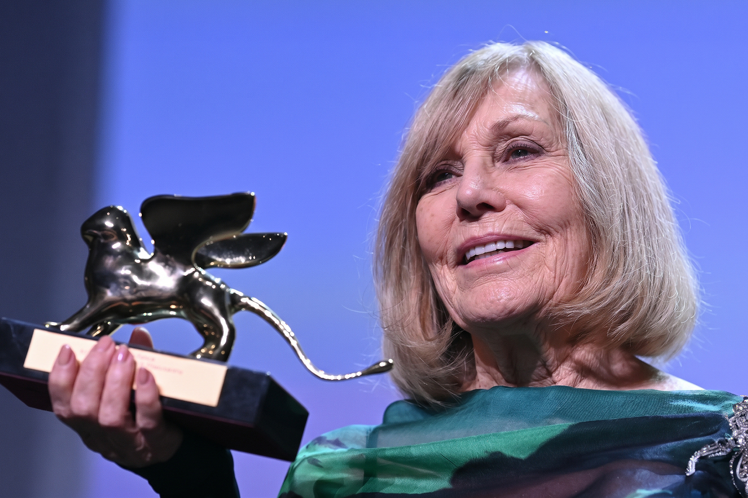 Kim Novak sostiene un trofeo del León de Oro y sonríe con su vestido verde y negro, mientras el premio refleja las luces del escenario sobre un fondo azul-púrpura. | Fuente: Gemini