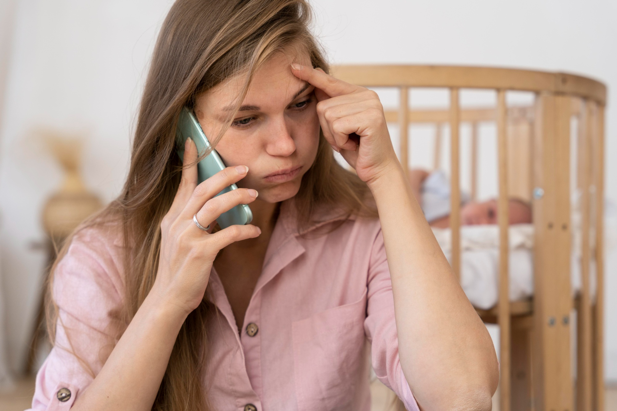 Una mujer frustrada hablando por teléfono con su bebé tumbado en una cuna detrás de ella | Fuente: Freepik