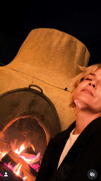 Selma Blair descansa con los ojos suavemente cerrados junto a una chimenea kiva tradicional de adobe encendida durante una reunión nocturna el 11 de octubre de 2026. La luz ámbar del fuego ilumina el rústico hogar curvo mientras ella se recuesta con un abrigo de polar oscuro sobre una camisa blanca con cuello. | Fuente: Instagram/selmablair