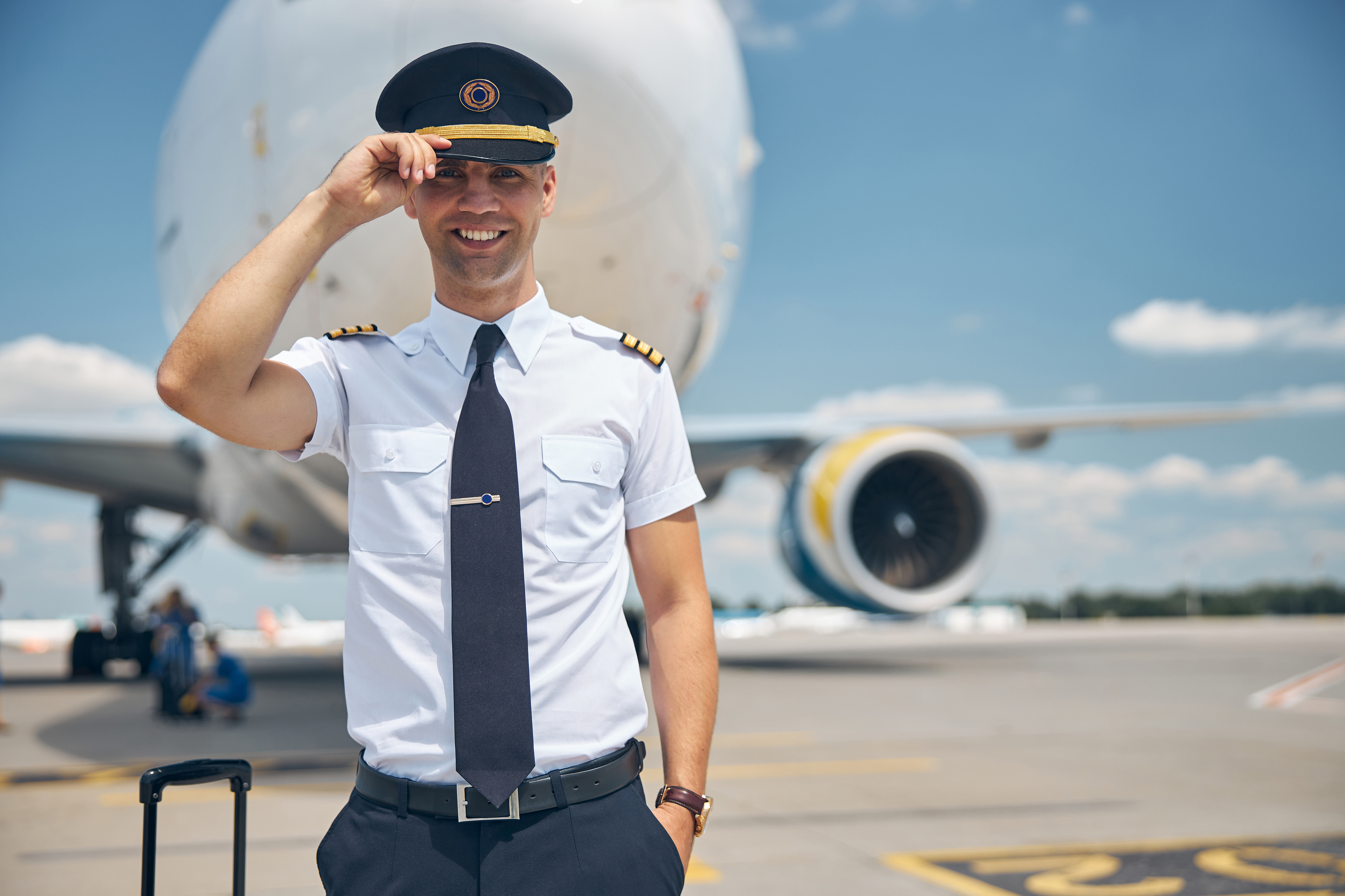 Alegre joven trabajador de aerolínea tocándose la gorra de capitán y sonriendo de pie en un aeródromo con un avión de fondo. | Fuente: Shutterstock