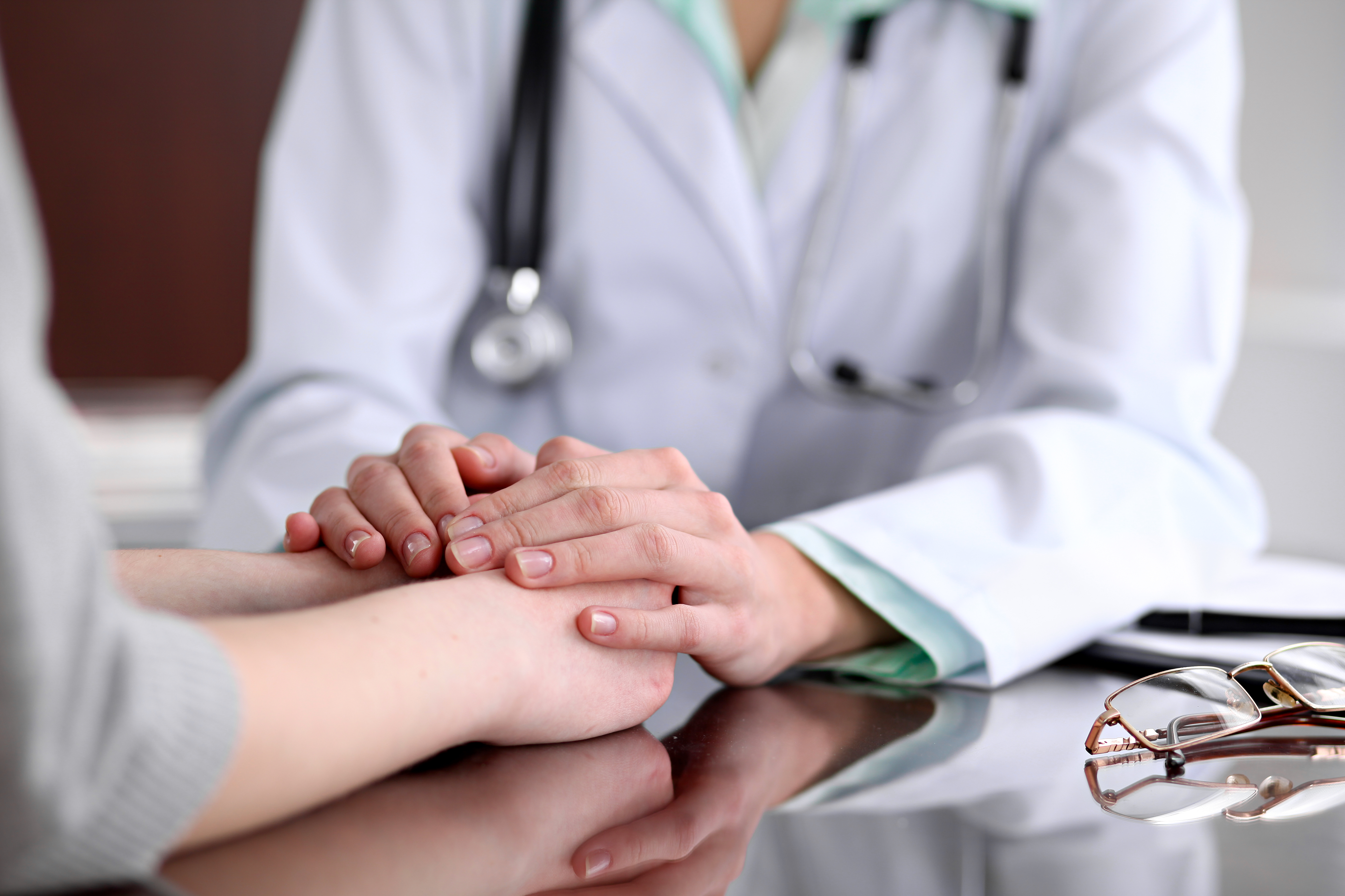 Amistosa doctora cogiendo de la mano a un paciente sentado en el escritorio para animarle, mostrarle empatía, animarle y apoyarle durante un examen médico. | Fuente: Shutterstock