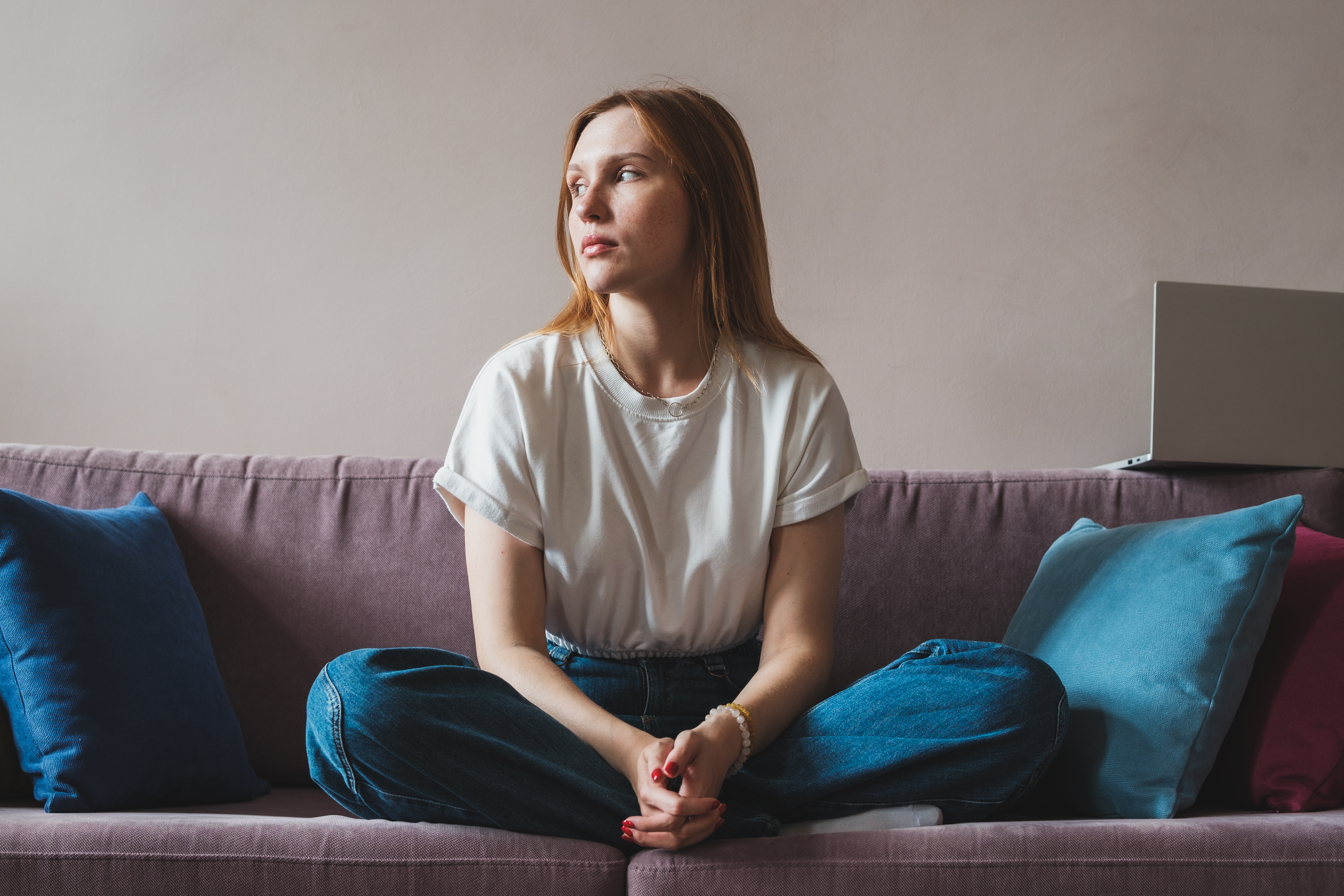 Joven autónoma deprimida y disgustada sentada en el sofá con un portátil mirando por la ventana | Foto: Getty Images