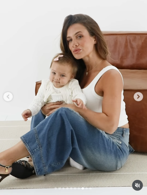 Sentada sobre el suelo frente a un sillón de cuero, Camila posó sonriente sosteniendo en su regazo a Aitana. La influencer combinó un top musculosa básico con pantalones de denim oxford y sandalias de tacón oscuro, mientras la pequeña lucía un delicado saquito blanco tejido y un moño a tono. | Fuente: Instagram/camihoms