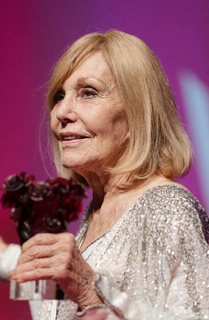 Kim Novak se gira ligeramente de perfil mientras sostiene un premio con motivos florales de color rojo oscuro; su chaqueta plateada brillante refleja la luz sobre un fondo rosa intenso. | Fuente: Gemini