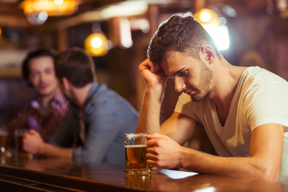Hombre con camiseta blanca mirando un vaso de cerveza mientras está sentado en la barra de un bar. | Imagen: Shutterstock