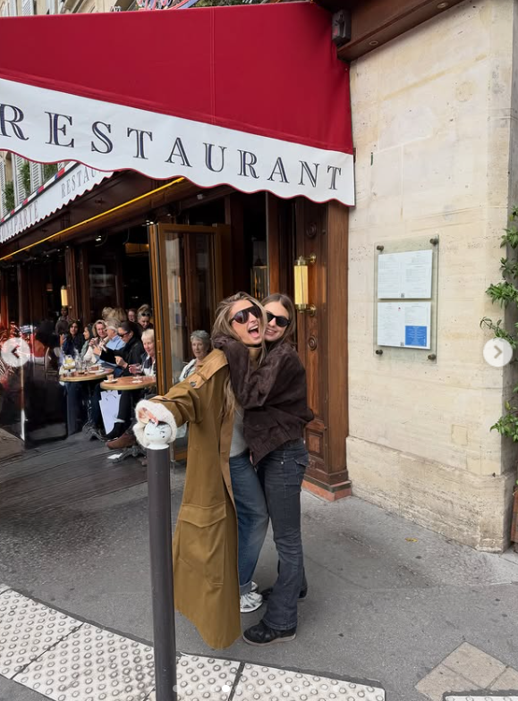 Valentina Barbier y su hermana Antonia Macri se abrazan con alegría y complicidad en la vereda frente a la fachada de un bistró parisino, ambas vestidas con abrigos de temporada, jeans holgados y anteojos de sol oscuros. | Fuente: Instagram/juliana.awada