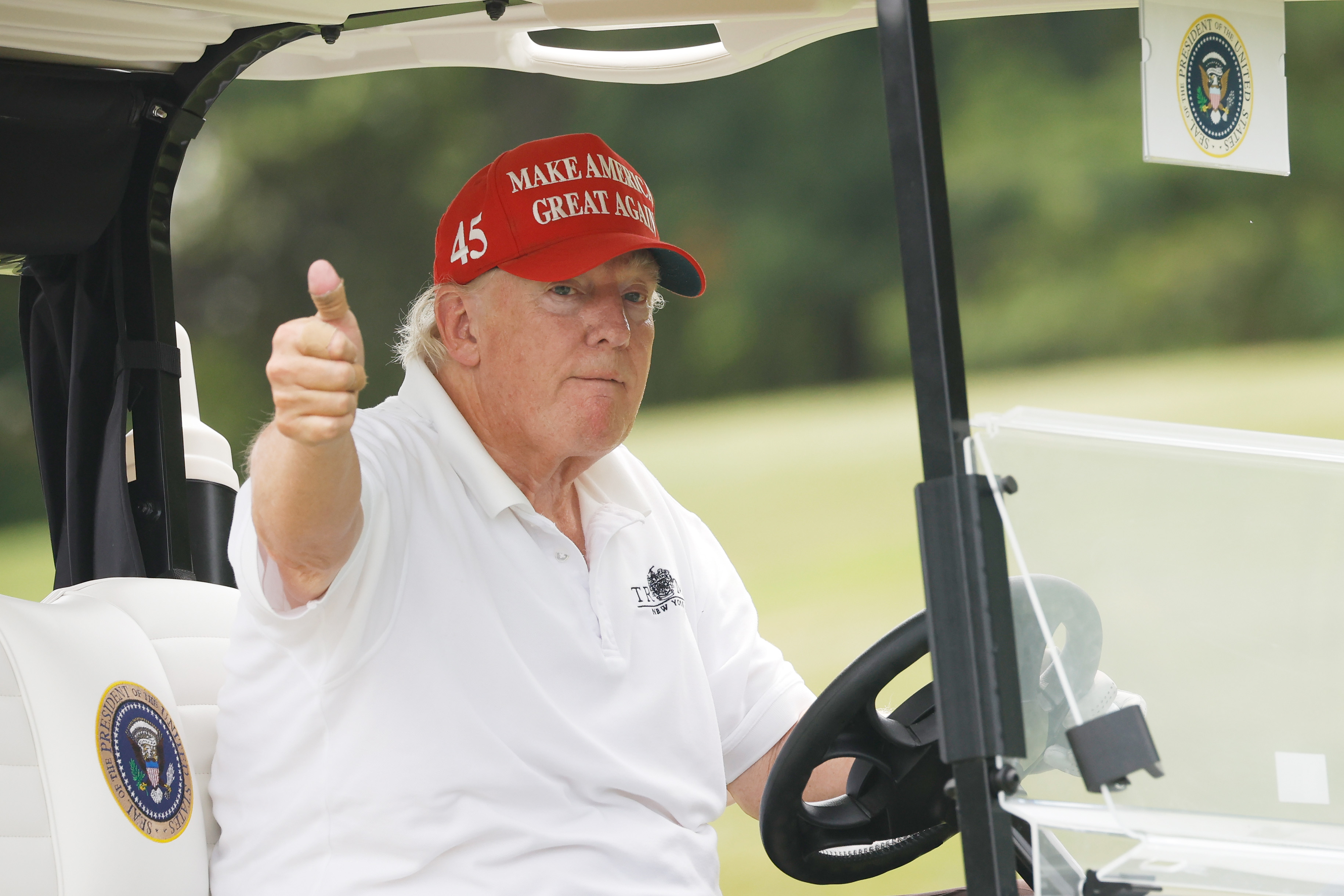 Sentado en un carrito de golf, Donald Trump levanta el pulgar con gesto serio. La diferencia de color entre su cara y su cuello es visible a la luz del día.