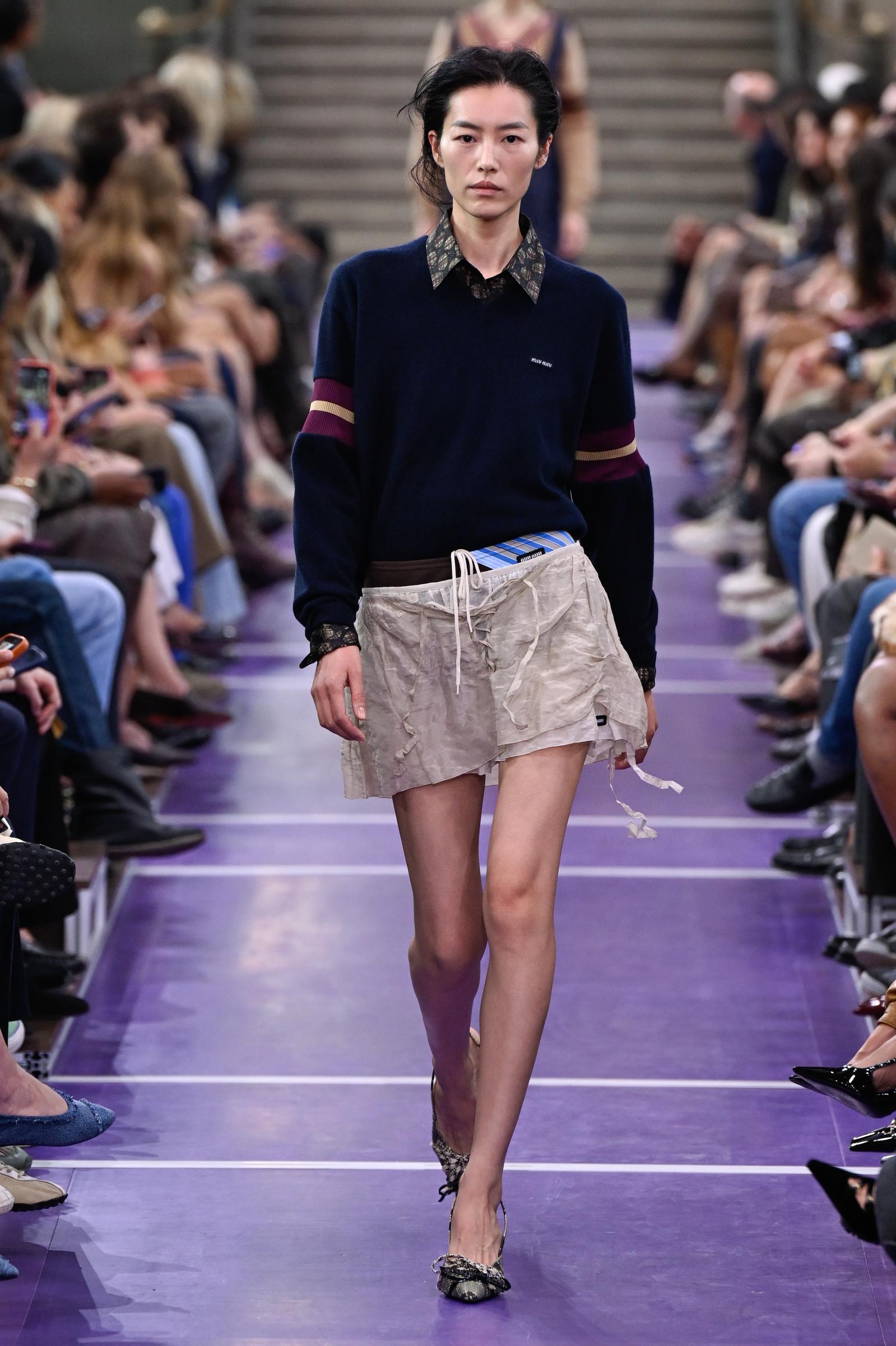 Liu Wen desfila por la pasarela de Miu Miu con un jersey azul marino con rayas burdeos y crema en las mangas, combinado con una camisa estampada y unos pantalones cortos holgados de color crema, y remata el look con unas bailarinas de punta. | Fuente: Getty Images