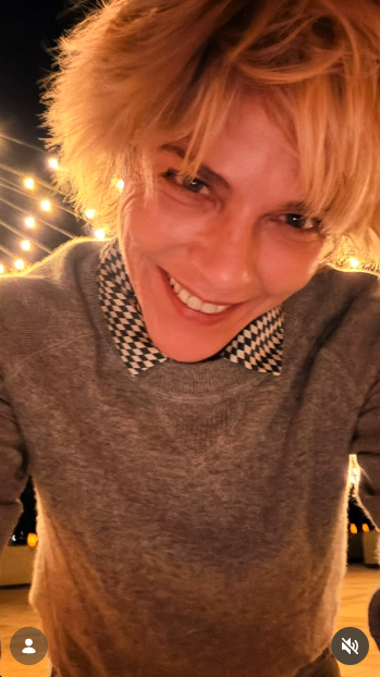 Sonriendo directamente a la cámara desde arriba, Selma Blair se hace un selfi nocturno bajo las luces resplandecientes de una cafetería el 11 de octubre de 2026. Lleva un jersey gris sobre un cuello con estampado de pata de gallo, y su flequillo rubio y con textura le cae sobre la frente. | Fuente: Instagram/selmablair