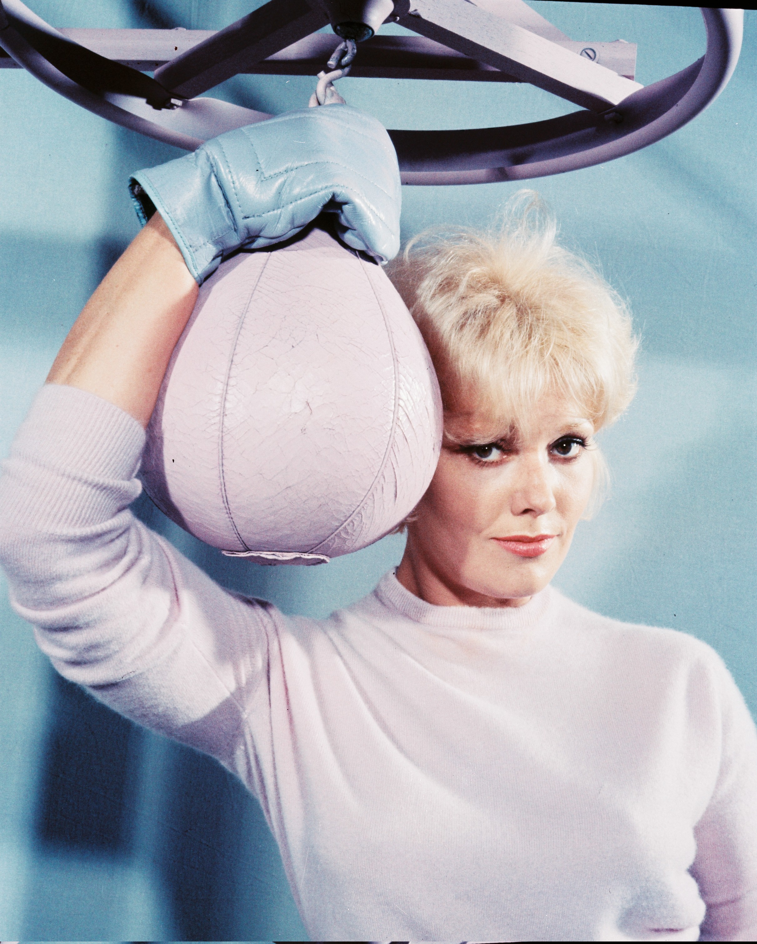 Kim Novak posa con un suéter blanco de lana y guantes de boxeo azules junto a un saco de boxeo colgado en un retrato de estudio. | Fuente: Getty Images